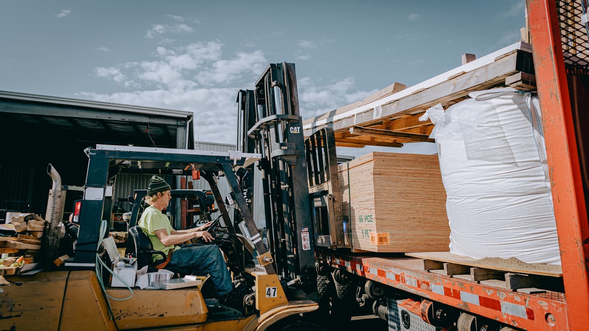 A forklift operator loading plywood onto a flatbed truck at a lumber yard