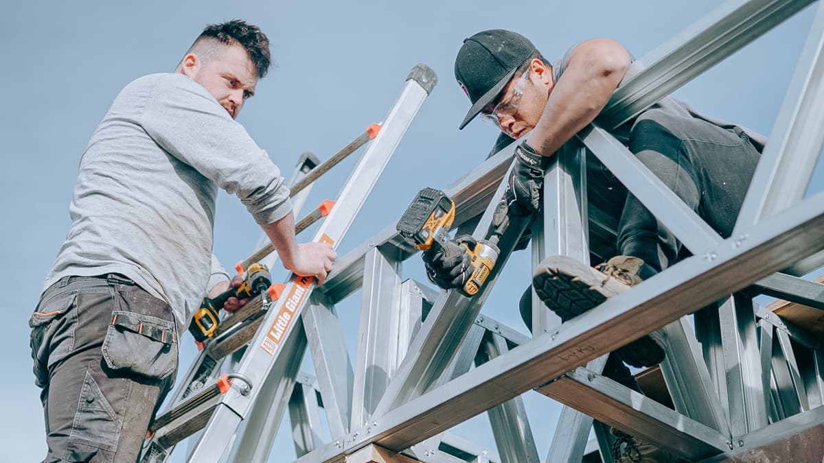 Two contractors on a jobsite drilling a metal frame together, one on a ladder