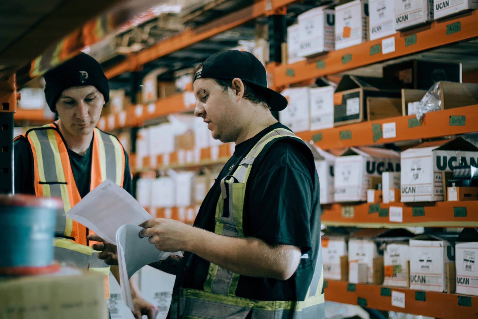 Two warehouse staff reading a printed material list in a racking aisle.